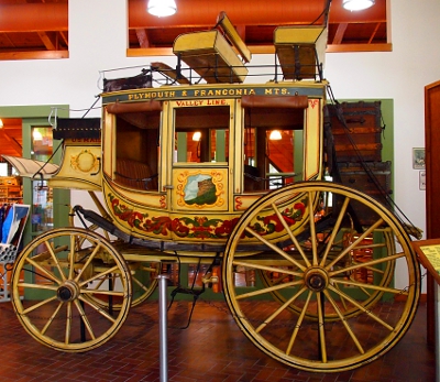 [A stage coach painted yellow-gold with seats on top as well as inside. Rear wheels twice the size of the front ones. Coach sits inside a museum.]